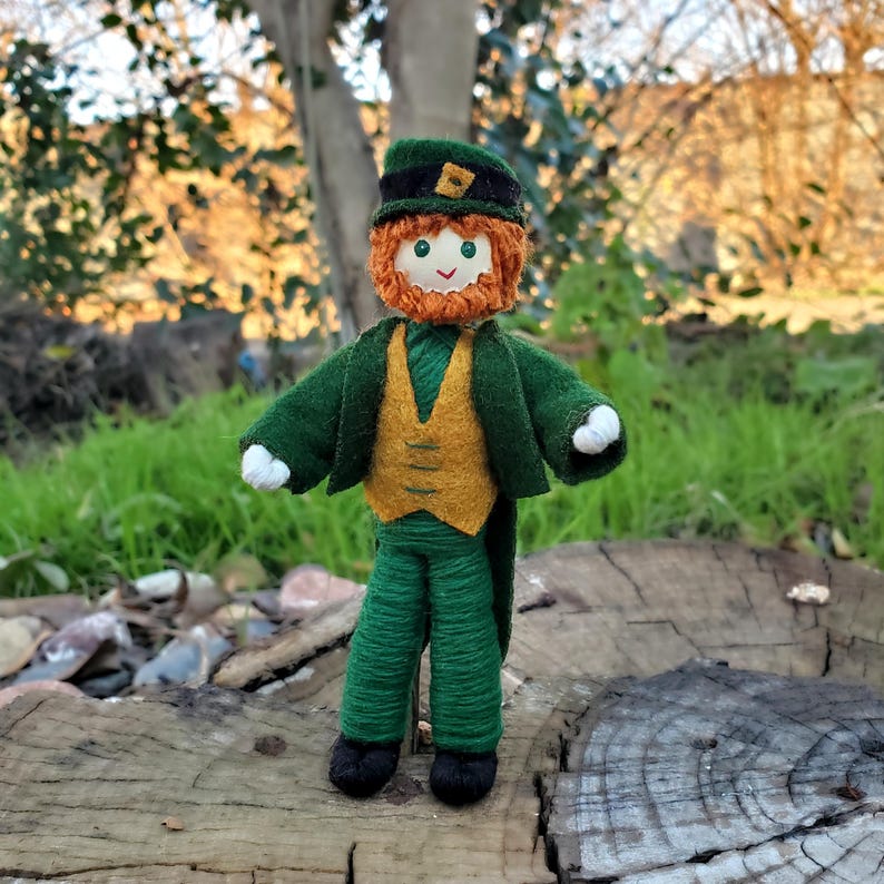 May include: A felt doll dressed as a leprechaun. The doll has a green jacket, a yellow vest, green pants, a green hat with a gold buckle, a red beard, and a friendly face.