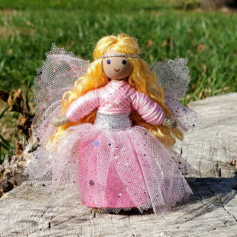 May include: A handmade doll with blonde yarn hair, a pink yarn dress, and a silver glitter belt. The doll is wearing a pink tulle skirt and has a silver glitter headband.