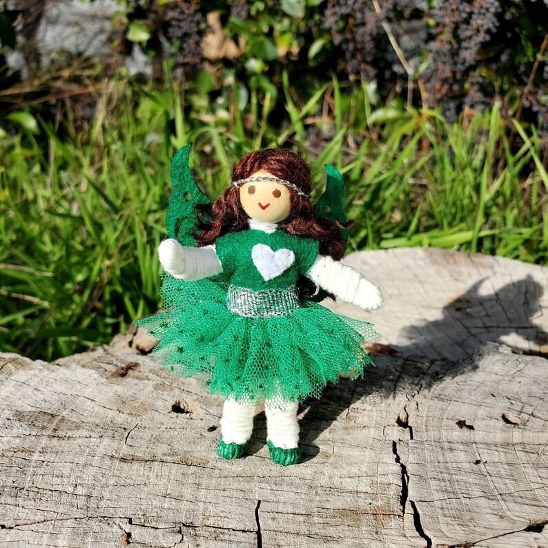 May include: A handmade fairy doll with green wings, a green dress, and a white heart on the front. The doll has brown hair and a painted face with a smile.
