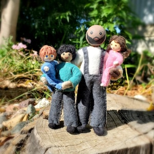 May include: A handmade felt family doll set. The dolls include a father figure in overalls, a child in a blue sweater, a child in a teal sweater, and a child in a pink outfit. The dolls are standing on a wooden surface with a natural background.