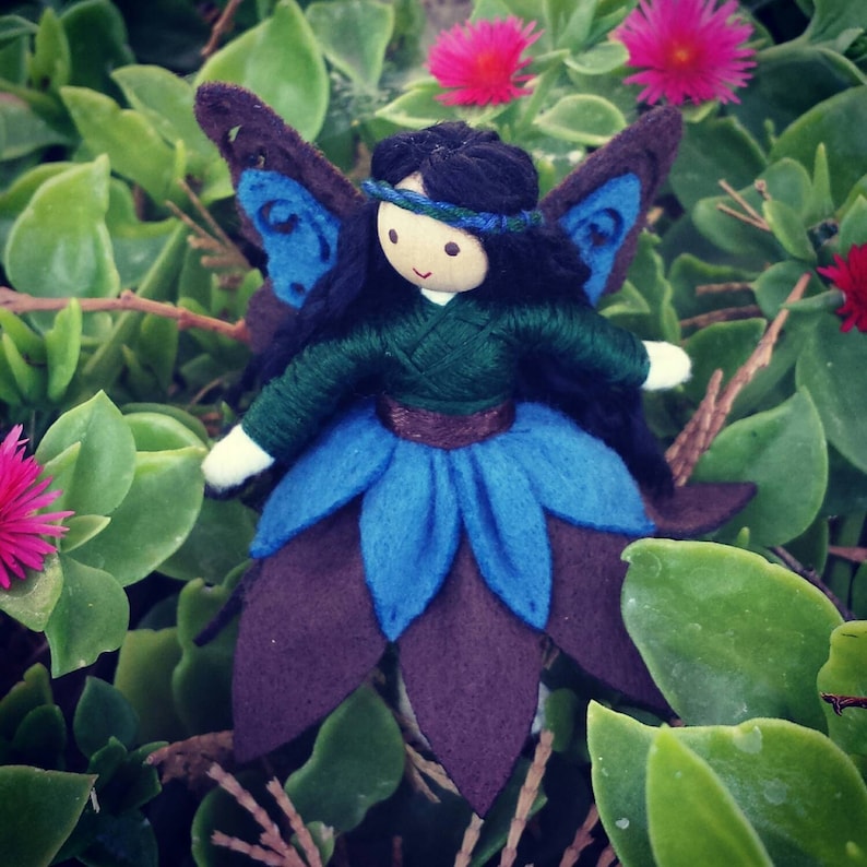 May include: A handmade felt fairy doll with blue wings, a green dress, and a brown flower skirt. The doll has black hair and a painted face.