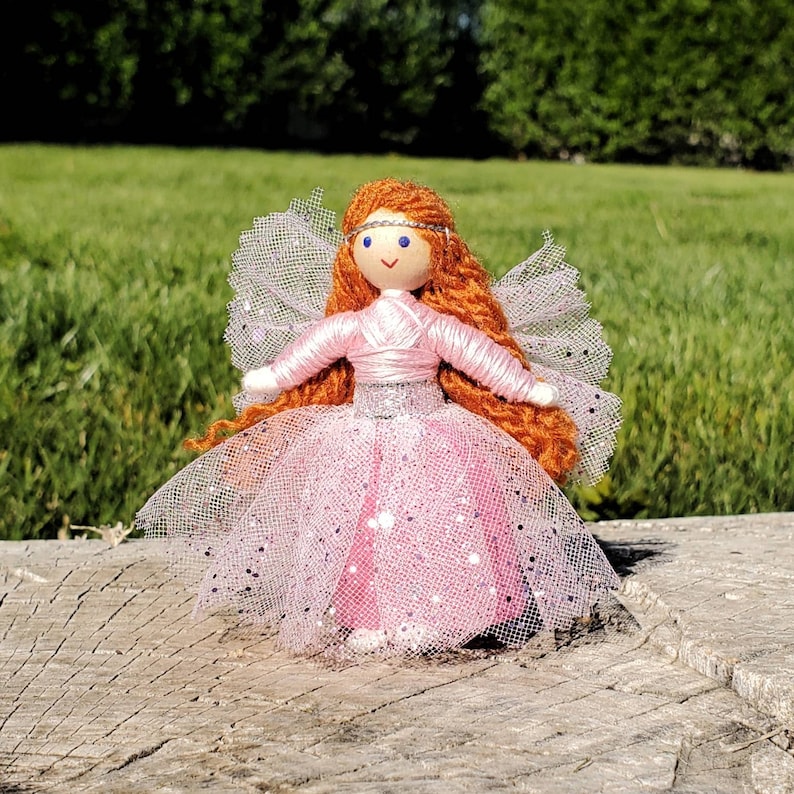 May include: A handmade doll with long, red yarn hair, wearing a pink tulle dress with silver glitter. The doll has a painted face with blue eyes and a pink crown.