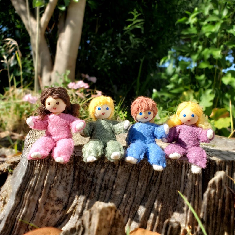 May include: Four handmade dolls with yarn hair and button eyes sit on a wooden surface. The dolls wear pink, green, blue, and purple outfits. The dolls are sitting in front of a green background.