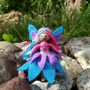 May include: A handmade fairy doll with pink yarn hair, a pink and purple dress, and blue wings. The doll is sitting on a rock.