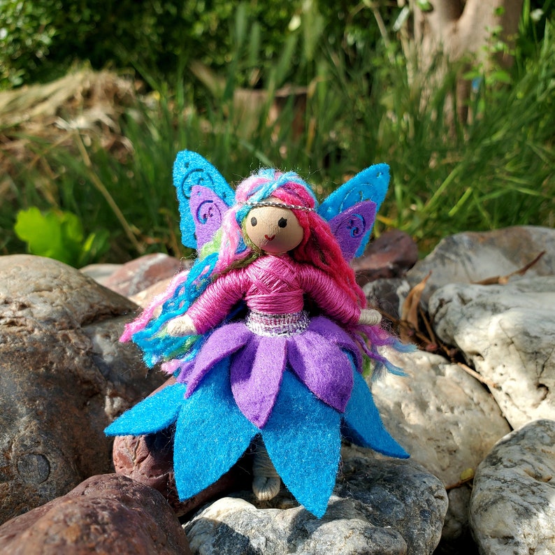 May include: A handmade fairy doll with pink yarn hair, a pink and purple dress, and blue wings. The doll is sitting on a rock.