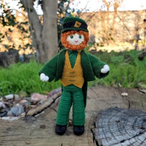 May include: A felt doll dressed as a leprechaun. The doll is wearing a green jacket, a gold vest, green pants, and a green hat with a gold buckle. The doll has a red beard and a smiling face.