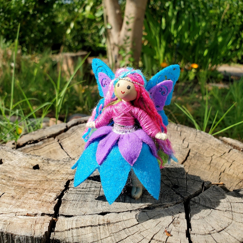 May include: A handmade felt fairy doll with pink yarn hair, blue and purple wings, and a blue and purple dress. The doll is sitting on a tree stump.