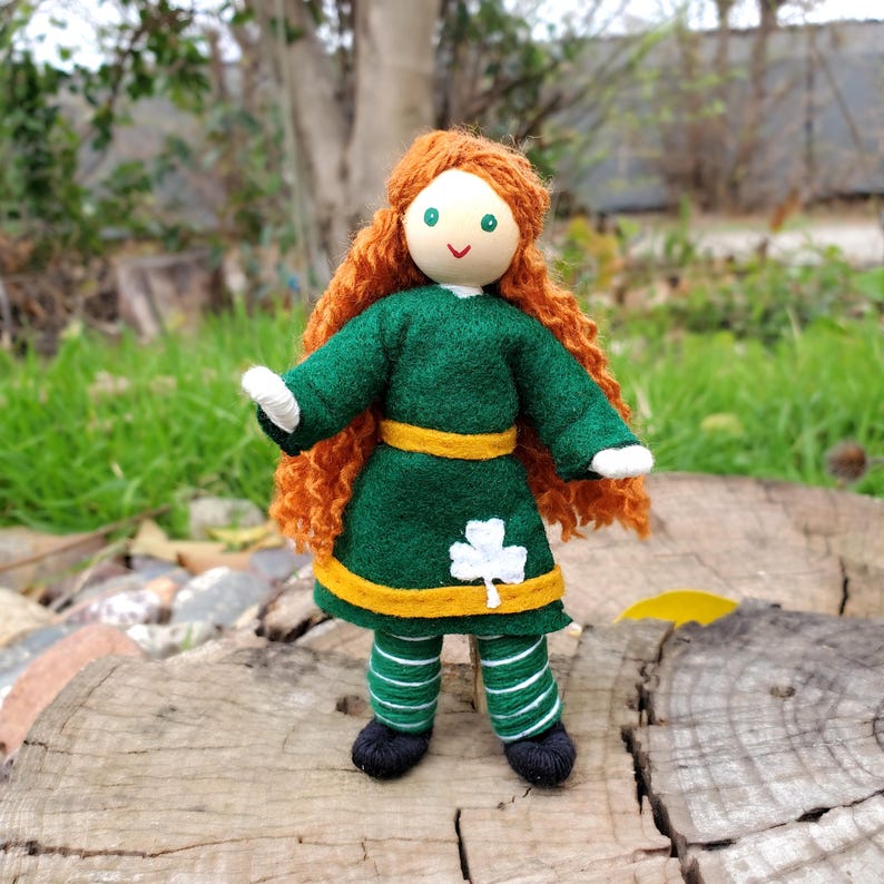 May include: A handmade doll with red curly hair, a green dress with a yellow sash and a white shamrock, and green and white striped pants.