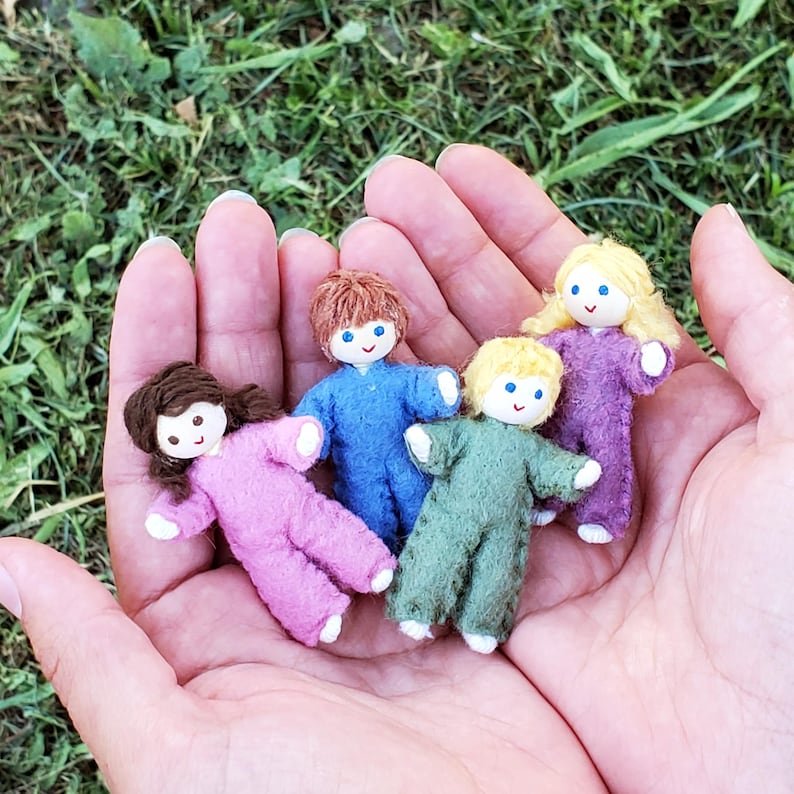 May include: Four small handmade dolls with felt bodies and yarn hair are held in an open hand. The dolls are dressed in pink, blue, green, and purple. Each doll has a painted face with blue eyes and a small smile. The dolls are a charming, whimsical craft.