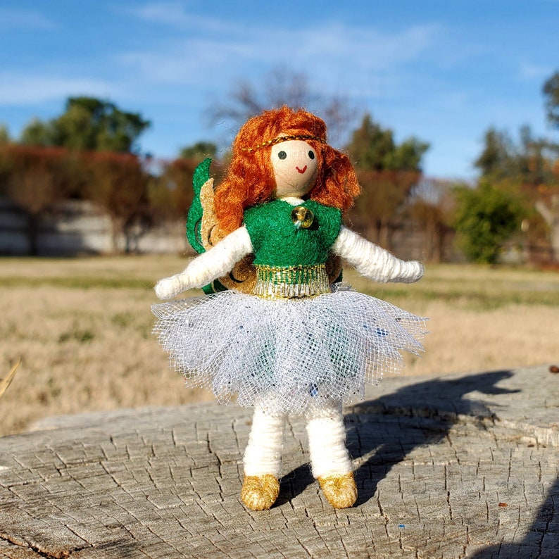 May include: A handmade doll with red hair, a green dress, and a white tulle skirt. The doll has a green and gold belt and a green and gold crown. The doll is standing on a wooden surface.