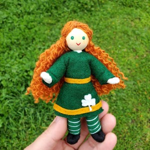 May include: A handmade doll with long, curly red hair, wearing a green dress with a yellow sash and a shamrock design. The doll has a white face with painted green eyes and a smile.