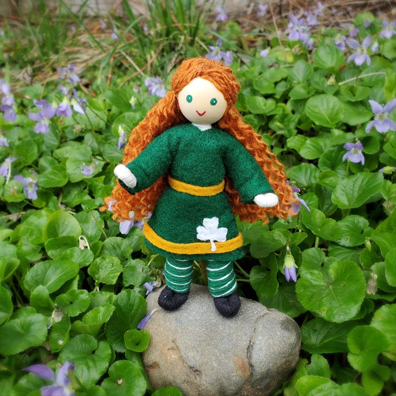 A handmade Irish doll with red hair, a green dress with a gold trim and a shamrock, and green and white striped pants. Girl leprechaun toy for Saint Patrick's Day.