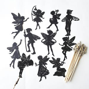 May include: A collection of ten black paper fairy silhouettes in various poses, including figures with wings, some holding staffs or flowers. Several wooden sticks with metal alligator clips are included for displaying the paper cutouts.