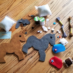 May include: A felt craft kit for making two horse figurines. The kit includes two felt horse shapes, one brown and one gray, along with felt pieces for the horse's mane, tail, and hooves. The kit also includes two spools of thread, stuffing, and a needle.