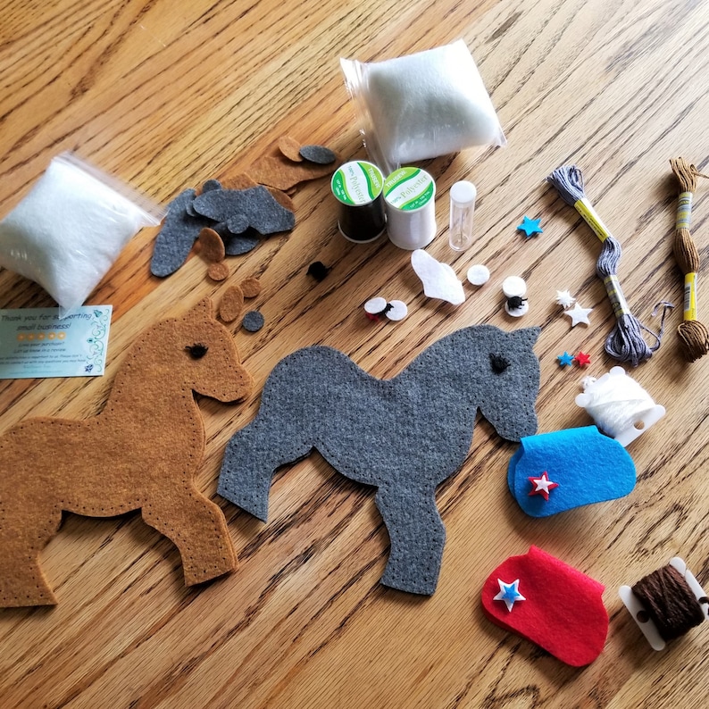 May include: A felt craft kit for making two horse figurines. The kit includes two felt horse shapes, one brown and one gray, along with felt pieces for the horse's mane, tail, and hooves. The kit also includes two spools of thread, stuffing, and a needle.
