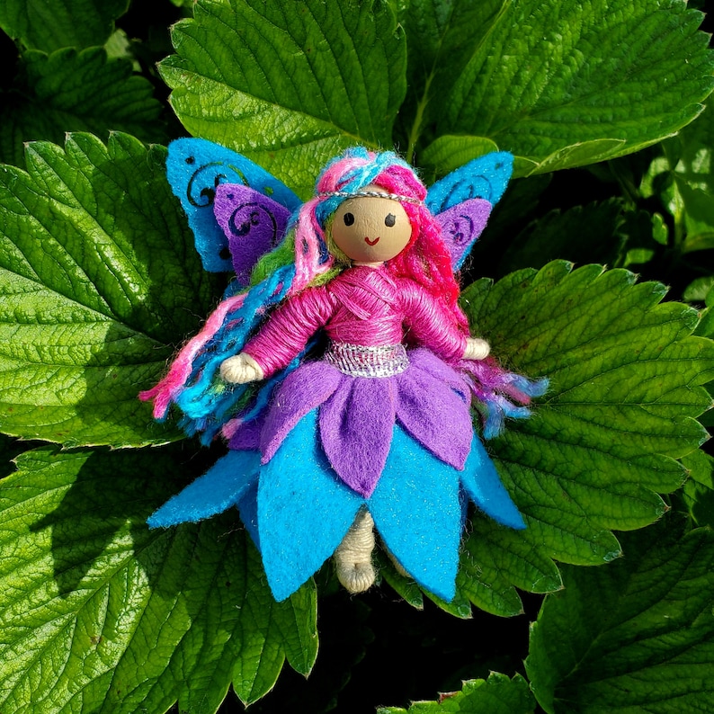 May include: A handmade doll with Rainbow yarn hair, a pink shirt, a purple and blue skirt and wings. The doll is wearing a silver belt and has a painted wooden face.