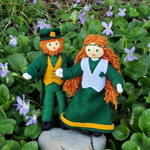 May include: A pair of handmade felt dolls dressed in green and gold, representing a leprechaun and a fairy. The leprechaun wears a green hat with a gold buckle and a green vest. The fairy wears a white vest with a shamrock design and a green dress with a gold trim.