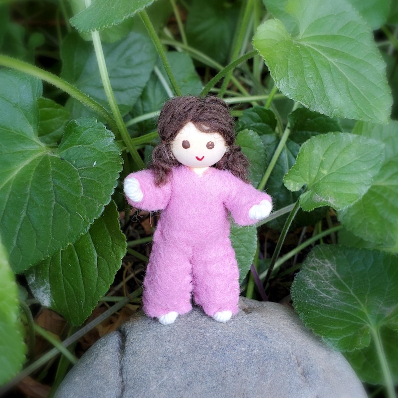 May include: A handmade doll with a painted face and brown yarn hair. The doll is wearing a pink jumpsuit with white accents on the hands and feet. It is standing on a gray rock, surrounded by green leaves.