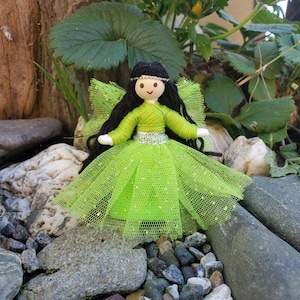 May include: A handmade fairy doll with black yarn hair, a green bodice, and a matching tulle skirt. The doll has white hands and a silver belt. The fairy has green glittery wings and a silver headband. The doll is sitting on a bed of rocks.