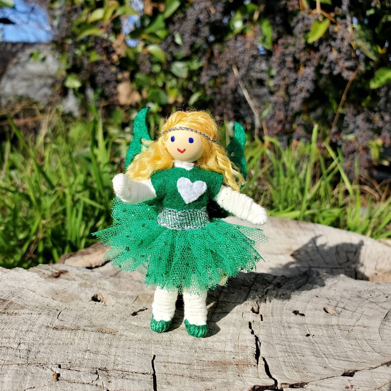 May include: A handmade doll with blonde hair, a green dress, and white wings. The doll is wearing a white heart on its chest and a silver belt. The doll is standing on a wooden surface.