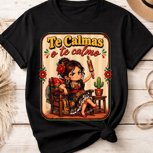 以下が含まれることがあります： 黒いTシャツ。女性が麺棒を持っている漫画のイラスト。テキスト「Te Calmas o te calmo」は黄色と緑色。女性は花柄のドレスと赤い靴を履いています。デザインはメキシコをテーマにしています。