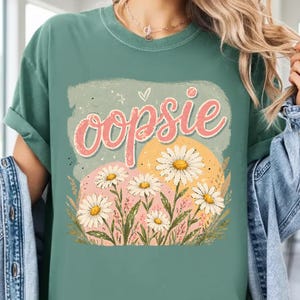 Puede incluir: Una camiseta verde con la palabra "oopsie" en escritura rosa. La camiseta presenta un diseño floral con margaritas y un fondo verde claro. El diseño incluye un corazón y estrellas.