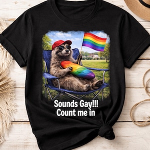 May include: Black t-shirt featuring a raccoon wearing sunglasses and a red cap, holding a rainbow flag and a rainbow-colored loaf of bread. The text "Sounds Gay!!! Count me in" is printed below the image.