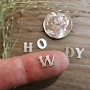 May include: Close-up of small, silver metal letters spelling "HOWDY" with a dime and sprigs of grass on a wooden surface. The letter "W" is held on a finger. The letters are approximately 0.5 inches in height.