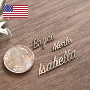 May include: Three silver name cutouts: "Bryce", "Merle", and "Isabella" in a cursive font. A U.S. dime is next to the names for size comparison. The names are on a wooden surface. An American flag is in the upper left corner.