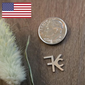 May include: A small, silver-colored pendant with the letters "76C" next to a U.S. dime. The dime is dated 2024. The pendant and coin are on a dark brown surface, with a sprig of dried plant matter in the background.