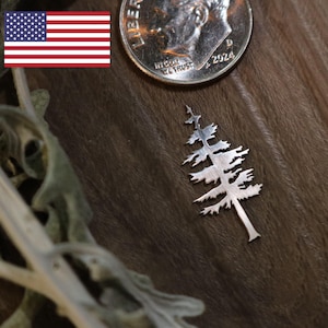 May include: A small, silver-colored tree pendant with detailed branches. A US quarter coin is next to the pendant for size comparison. The pendant is on a brown surface.