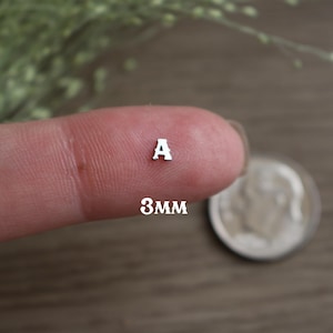 May include: A tiny silver letter "A" is displayed on a fingertip, with the text "3mm" below it. A silver coin is visible in the background. The image is taken in a close-up shot.