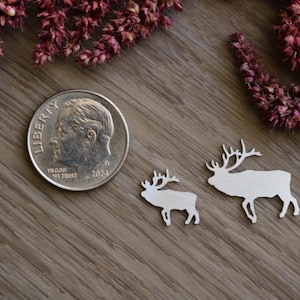 May include: Two silver-colored metal elk-shaped cutouts positioned next to a United States ten-cent coin for scale. The elk shapes feature detailed antlers and are rendered in a flat, metallic finish, showing a clear size comparison to the coin.