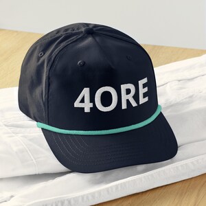 Puede incluir: Gorra azul marino con la palabra "4ORE" bordada en blanco. La gorra presenta un detalle de cuerda verde turquesa a lo largo del borde. La gorra está sobre una superficie blanca.