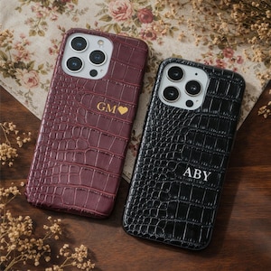 Peut inclure: Deux coques de téléphone avec une texture peau de crocodile. L'une est bordeaux avec les initiales "GM" et un cœur, l'autre est noire avec les initiales "ABY". Les coques sont posées sur un tissu à motifs floraux.