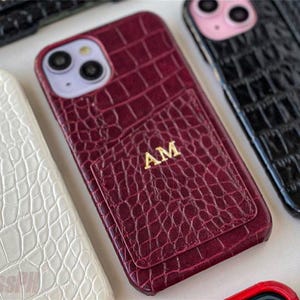 Puede incluir: Una colección de fundas personalizadas para teléfonos en varios colores. Las fundas presentan un patrón texturizado similar al de la piel de cocodrilo. Una funda es de color burdeos con las iniciales "AM" en dorado. Otras fundas son blancas, negras y rojas.