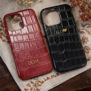 Può includere: Due custodie per telefono con texture a pelle di coccodrillo. Una è bordeaux con il nome "DOM" in lettere dorate. L'altra è nera con le iniziali "G.D" e un simbolo a cuore dorato. Entrambe le custodie hanno forma rettangolare.