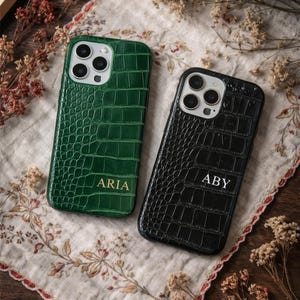 May include: Two phone cases with a crocodile skin texture. One is dark green with the name "ARIA" in gold lettering, and the other is black with the initials "ABY". The cases are on a floral embroidered cloth.