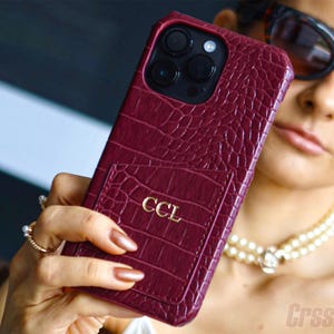 Puede incluir: Una funda de teléfono burdeos con textura de cocodrilo y tarjetero. La funda tiene las letras doradas "CCL" en el tarjetero. La funda del teléfono la sostiene una persona con gafas de sol y un collar de perlas.