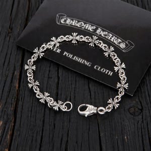 May include: A silver bracelet with a cross design and clasp. The bracelet is displayed on a black surface with a card that reads "CHROME HEARTS" and "SILVER POLISHING CLOTH".