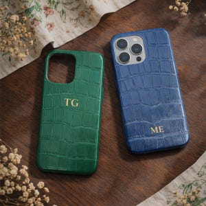 Può includere: Due custodie per telefono con texture a pelle di coccodrillo. Una è verde con le iniziali "TG" in oro, l'altra è blu con le iniziali "ME" in oro. Le custodie sono su una superficie di legno con tessuto floreale.