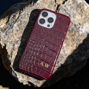 May include: A burgundy phone case with a crocodile skin texture. The case has a glossy finish and features a camera cutout for multiple lenses. Gold initials "A.W" are visible on the lower part of the case.