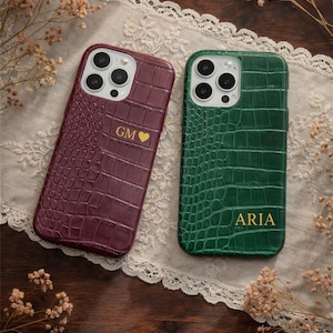 Può includere: Due custodie per telefono con texture a pelle di coccodrillo. Una è bordeaux con "GM" e un cuore, l'altra è verde con "ARIA". Entrambe le custodie sono su un runner di pizzo bianco con fiori secchi.