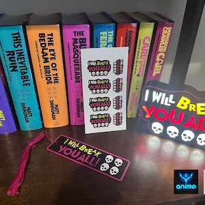 Könnte beinhalten: Eine Sammlung von Büchern mit farbenfrohen Buchrücken, darunter Titel wie "A Parade of Horribles" und "This Inevitable Ruin." Ein Lesezeichen und ein Leuchtschild mit dem Text "I Will Break You All!" sind ebenfalls zu sehen. Aufkleber mit Totenkopf-Grafiken sind vorhanden.