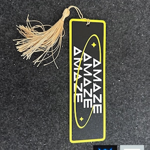 May include: Black rectangular tag with the word "AMAZE" repeated three times in white, with a yellow border and design. A gold tassel hangs from the top. The tag also has the "animo" logo and "Made in the USA" label.