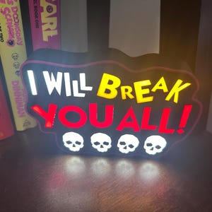 Könnte beinhalten: Ein beleuchtetes Schild mit dem Text "I WILL BREAK YOU ALL!" in Weiß, Gelb und Rot. Das Schild hat einen schwarzen Rand und vier weiße Totenkopfmotive. Bücher sind im Hintergrund sichtbar.