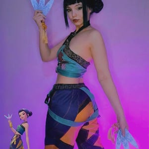 May include: A teal and gold halter top and color-blocked pants with orange and blue panels. The outfit is accessorized with blue, fan-shaped props. The text "miezute" is visible on the pants.
