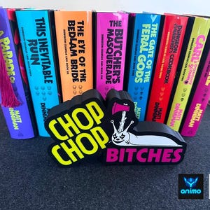 Könnte beinhalten: Eine Sammlung von acht Dungeon Crawler Carl-Büchern mit bunten Buchrücken, darunter Titel wie This Inevitable Ruin und The Butcher's Masquerade, sowie ein schwarzes Schild mit einer weißen Schneckengrafik und dem Text CHOP CHOP BITCHES.