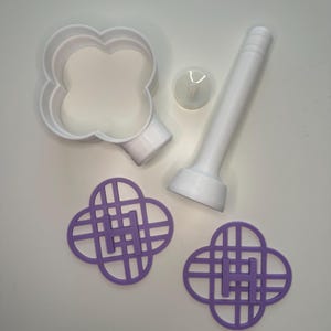May include: A white four-leaf clover-shaped cookie cutter with a handle, a small white round object, and two purple clover-shaped stencils with a lattice design. The items are arranged on a white surface.