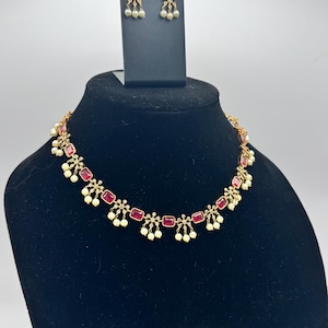 May include: A gold-toned necklace and earring set displayed on a black bust. The necklace and earrings feature rectangular red gemstones and small white pearls. The jewelry is designed with a delicate, ornate style.